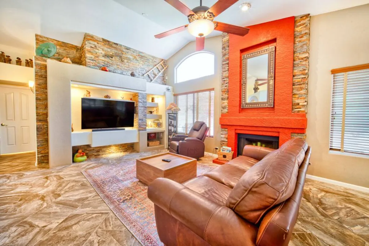 A warm living room centered around a stone fireplace, featuring a large television, a red accent wall, and plush, comfortable seating. The arrangement is conducive to both relaxing alone and gathering with friends or family for movie nights or casual conversations.