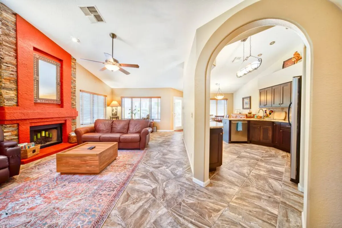 This image features a warm living area with a red accent wall, a stone fireplace, a leather sofa, and a wood coffee table. The space extends towards an adjacent kitchen, suggesting a seamless transition for gatherings and family activities.