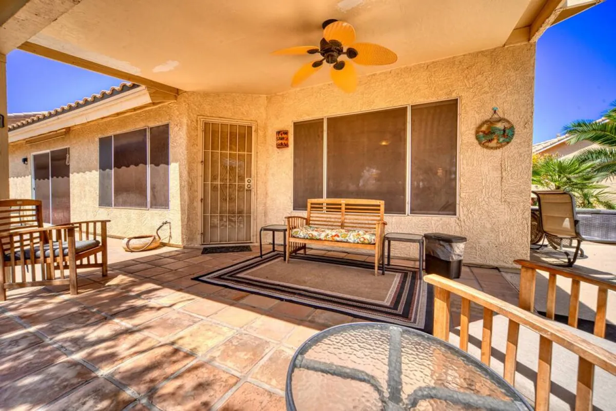 A comfortable and stylish patio outfitted with wooden furniture, a decorative ceiling fan, a colorful couch cushion, and a glass table. The scene is casual and open, ideal for relaxing outdoors, enjoying fresh air, or socializing after a day out.