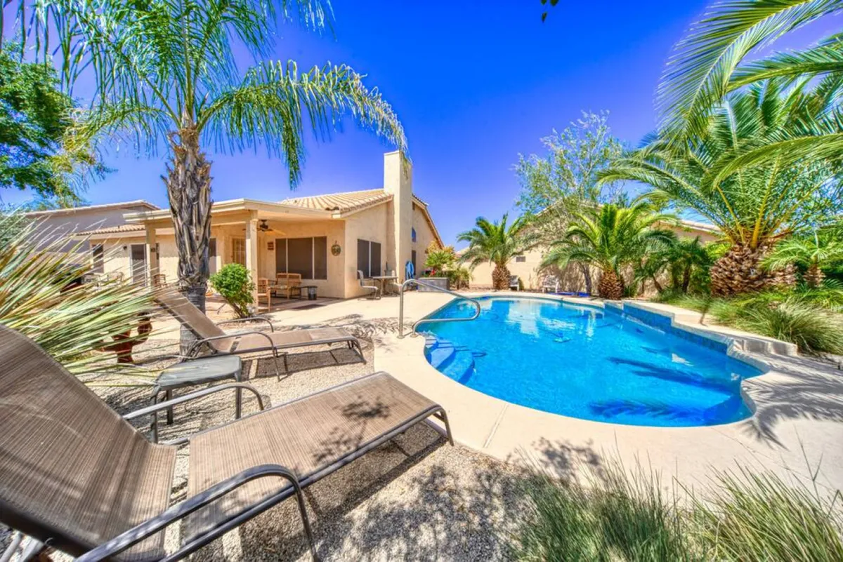 An inviting backyard view with the swimming pool framed by palm trees and lounge chairs. The patio area is also visible, showing more space for sunbathing or outdoor dining. The overall environment is bright, open, and exudes privacy and relaxation.