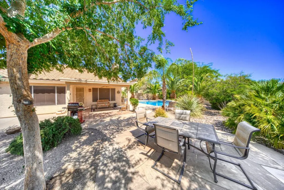 A serene backyard scene featuring a swimming pool, lush greenery, and a patio area with an outdoor dining table. The setting is relaxed and private, offering both opportunities for swimming and comfortable outdoor meals or quiet moments amidst nature.