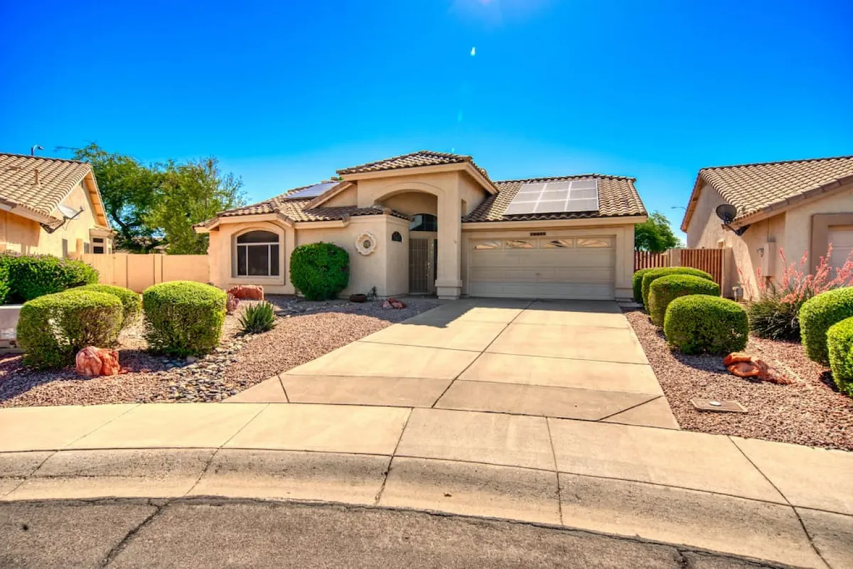 A view of the home's front yard with manicured landscaping, solar panels on the roof, and the garage entrance visible. The scene suggests a peaceful, sunny neighborhood setting, ideal for relaxed, long-term living.