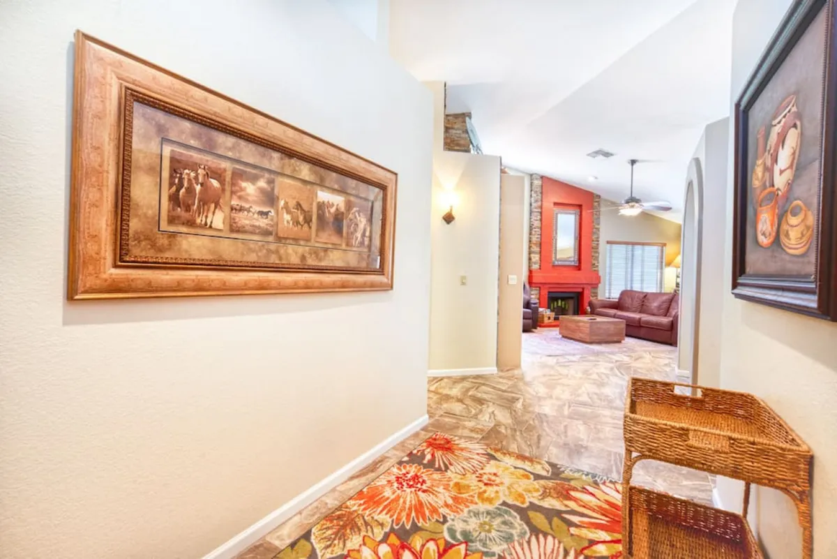 The image shows an open living area with a stone fireplace, a colorful floral rug, and framed artwork featuring horses. The view extends into a hallway, emphasizing the spaciousness and suitability for larger family gatherings.